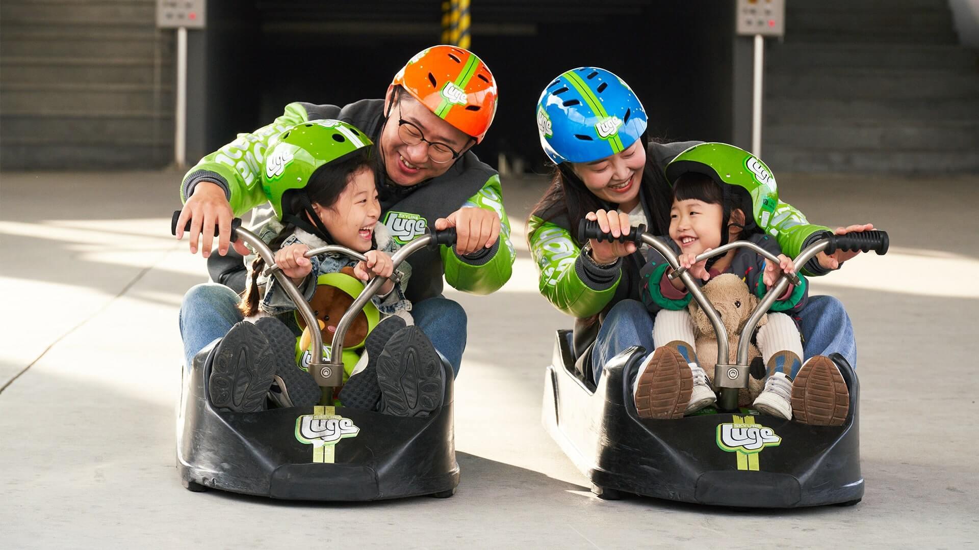 Two adults prepare to ride the Luge with small children in the same carts with them.