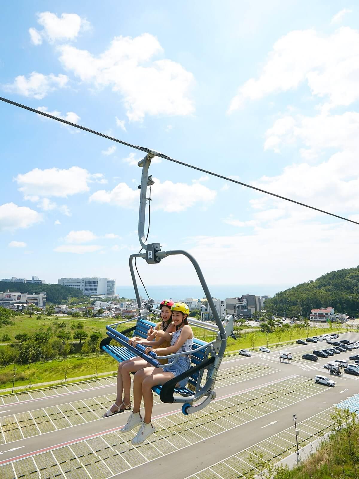 Two ladies ride the Skyline Luge Busan chairlift to the top station with panoramic views of the ocean behind.