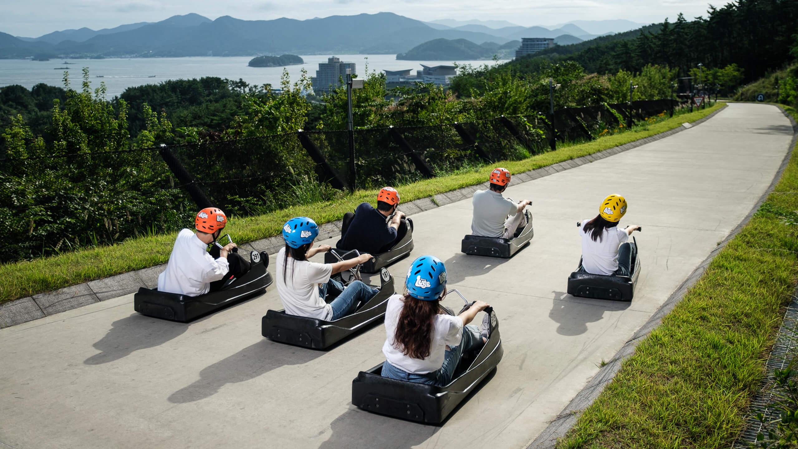 A group of people head down the Luge tracks together with views of Tongyeong city and the ocean behind them.