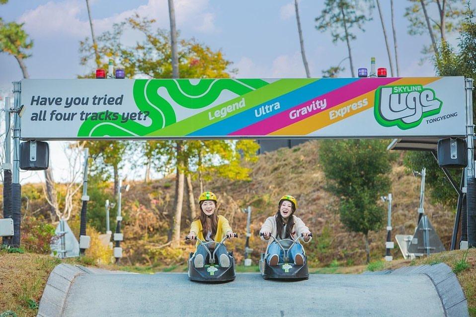 Two girl friends ride underneath a large banner showing off the different Luge tracks at Skyline Luge Tongyeong.